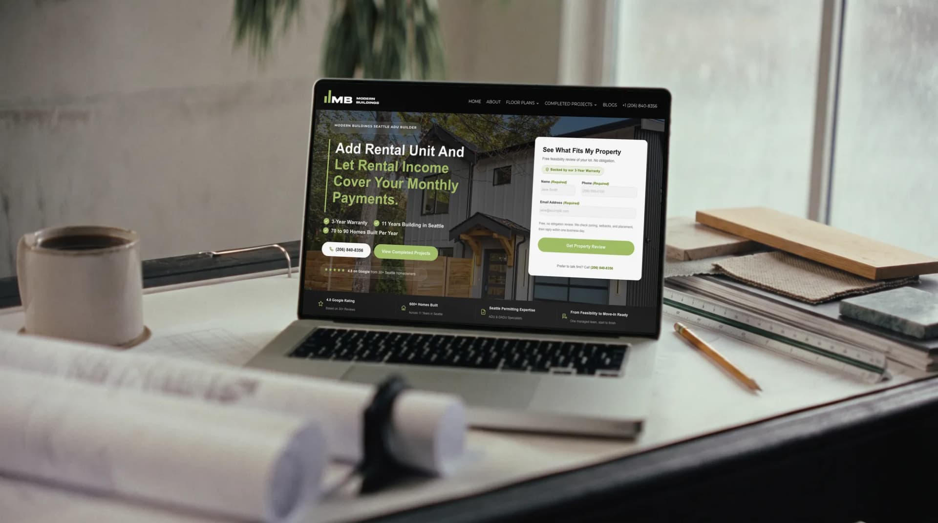 The Modern Buildings website open on a laptop on a drafting table, rolled plans beside it
