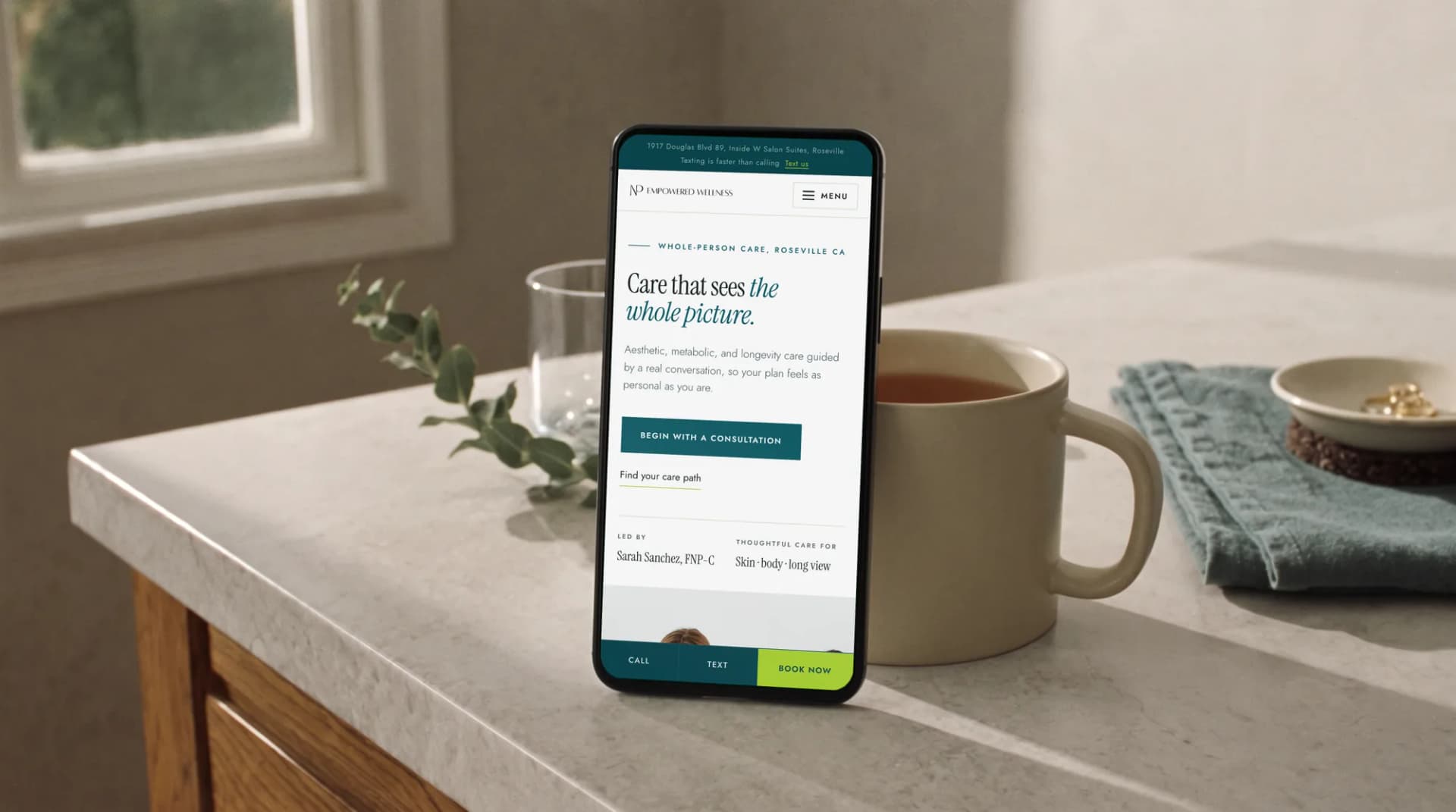 The NP Empowered Wellness website on a phone leaning on a mug on a sunlit kitchen counter